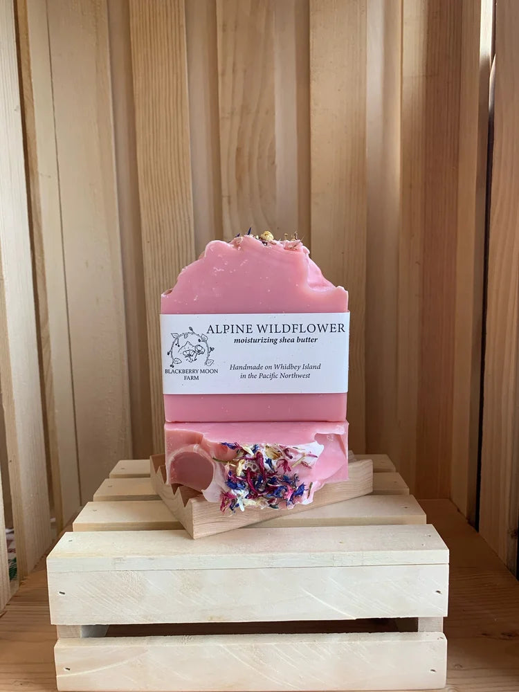 Alpine Wildflower handmade soap bars with pink color and dried flower pieces displayed on a wooden crate stand. 